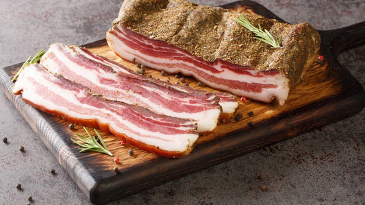 Raw peppered bacon strips on a wooden cutting board, with a piece of bacon in the background.