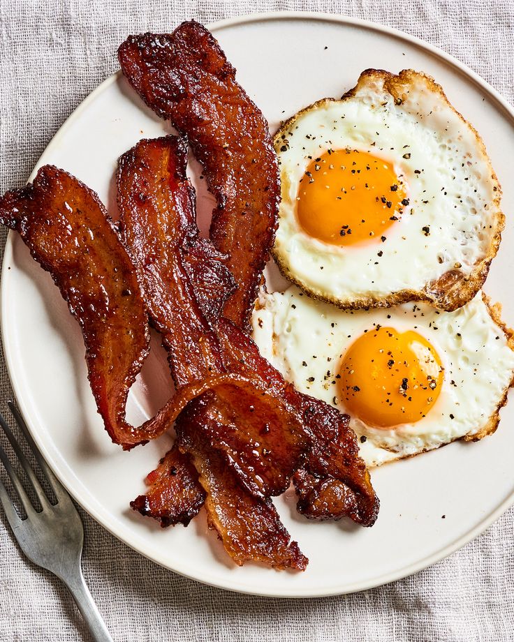 A plate containing roasted bacon and two fried eggs with a visible fork, set on a cloth surface.